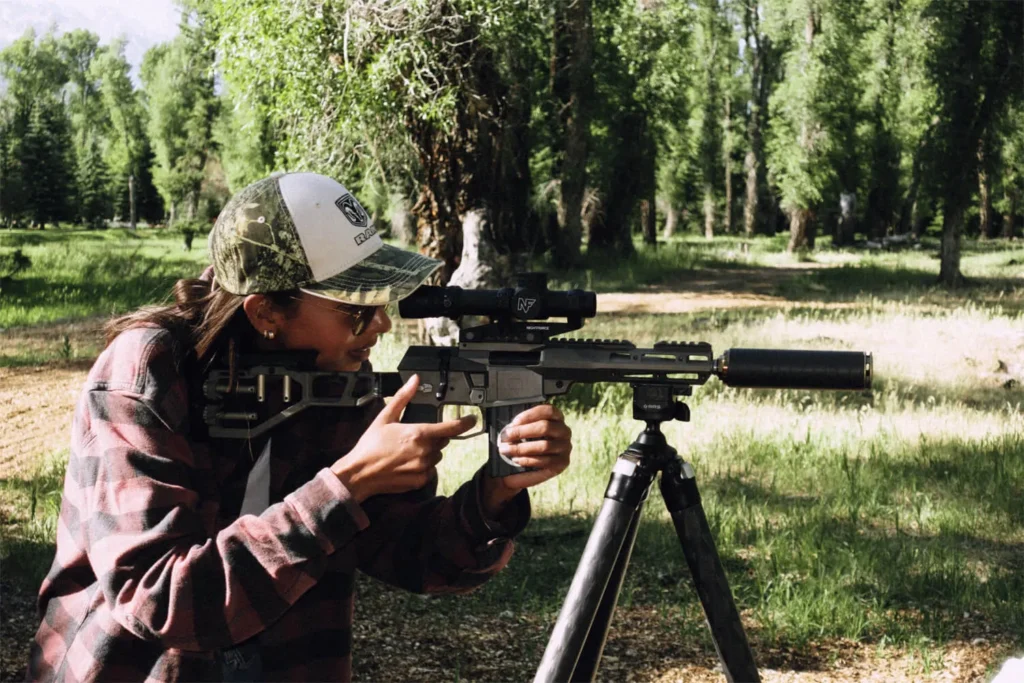 woman outdoors in the woods looking through a riflescop as she is shooting a suppressed AR rifle on a tripod. The gun is suppressed with a SilencerCo Scythe Ti.