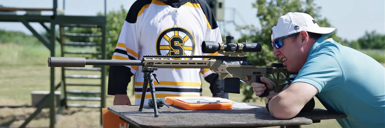man shooting a Daniel Defense Delta 5 Pro bolt action rifle in 6mm Creedmoor, suppressed with a SilencerCo Scythe-Ti, mounted with Bravo Direct Thread mount and single port Scythe Anchor Brake. A man stands in the background wearing a Happy Gilmore hockey jersey. This is from SilencerCo's 2nd annual Chubbs Peterson Rifle Golf and Hog Elimination tournament.